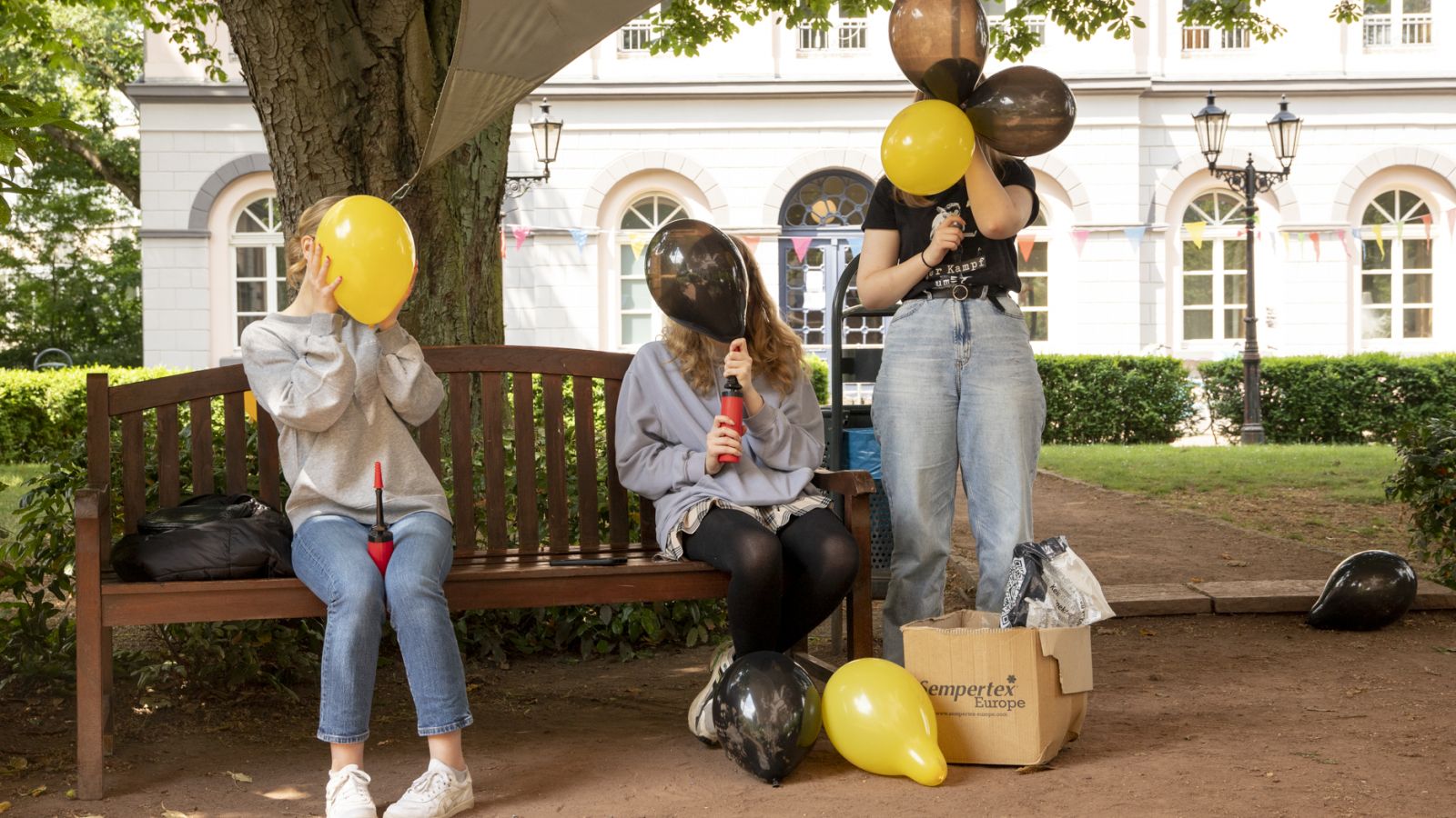 Junge Volunteers sitzen auf einer Parkbank unter einem Baum und blasen schwarze und gelbe Luftballons auf. Im Hintergrund ist das Marinsstift in Moers zu sehen.